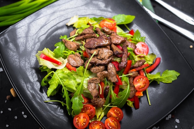 trozos de carne fritos, sobre verduras con tomates cherry, sobre un plato negro, sartén con verduras