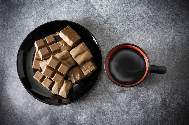 Trozos de chocolate diferente en un plato negro y una taza de café con vapor en la vista de la mesa gris arriba