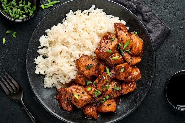 Trozos de filete de pollo teriyaki picante con arroz, cebollas verdes y semillas de sésamo negro en placa negra sobre una superficie de hormigón oscuro