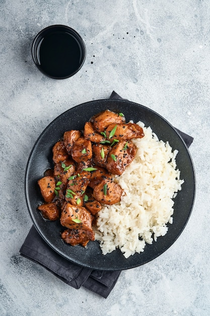 Trozos de filete de pollo teriyaki picante con arroz, cebollas verdes y semillas de sésamo negro sobre placa negra