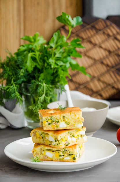 Trozos de tarta casera con huevos, cebollas verdes y atún enlatado en un plato blanco sobre un fondo de hormigón oscuro. Orientación vertical