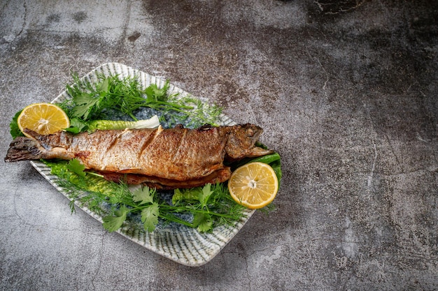 Trucha entera asada con limón, lechuga y eneldo y perejil en un plato contra una mesa de piedra gris