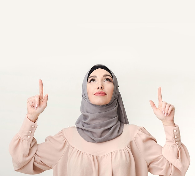 Tu anuncio aquí. Mujer en hijab apuntando hacia arriba en el espacio en blanco en la pared blanca.