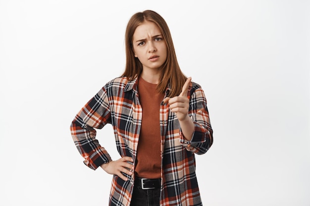 Foto tú mejor no. mujer joven seria sacudiendo el dedo y las cejas fruncidas disgustada, regañando el mal comportamiento, prohibir algo, prohibir o desaprobar, de pie contra el fondo blanco