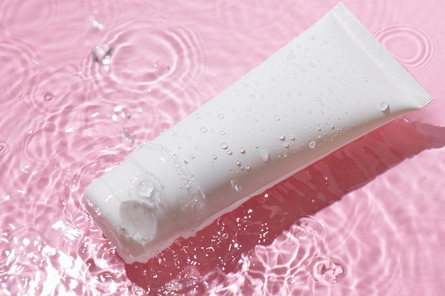 Tubo blanco en blanco de crema en salpicaduras de agua sobre fondo rosa Presentación de nuevo producto del concepto de cosméticos de belleza