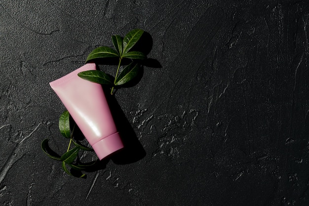 Tubo cosmético rosa con crema facial, loción corporal o limpiador sobre un fondo oscuro con hojas verdes frescas. Concepto de cuidado de la piel sensible. Copie el espacio, simule.