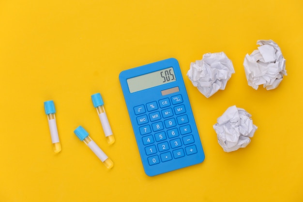 Tubos de ensaio, calculadora com bolas de papel amassadas em fundo amarelo. Vista do topo