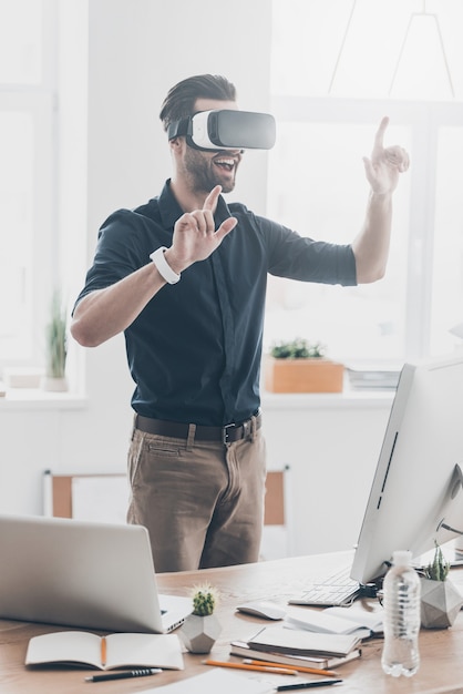 Tudo é tão real! Jovem bonito com fone de ouvido VR gesticulando e sorrindo enquanto está no escritório de criação