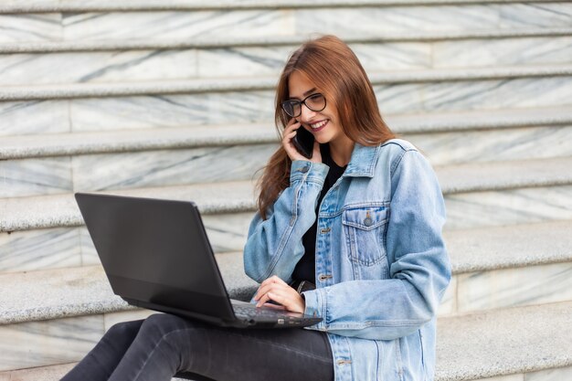 Tudo nos negócios. Jovem mulher elegante em uma jaqueta jeans e óculos usa um laptop e falando ao telefone enquanto está sentado na escada da cidade. Trabalho remoto.