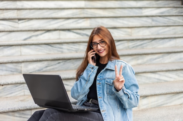 Tudo nos negócios. Jovem mulher elegante em uma jaqueta jeans e óculos usa um laptop e falando ao telefone enquanto está sentado na escada da cidade. Trabalho remoto.