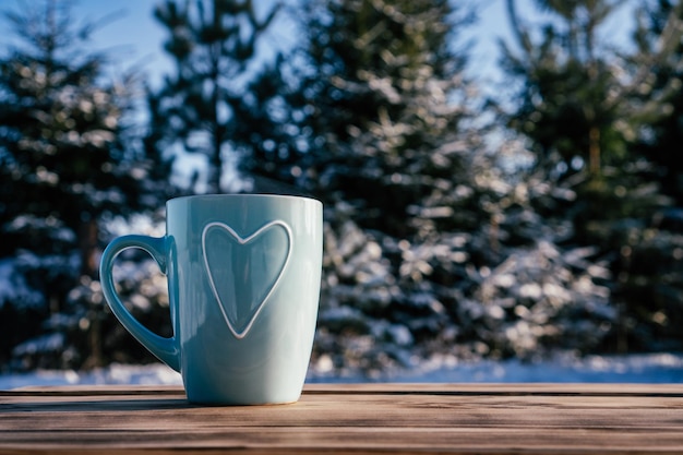 Türkisblaue Tasse mit aufsteigendem Herzdampf auf Holztisch vor dem Hintergrund von Kiefern und Tannen mit Schnee am Wintertag
