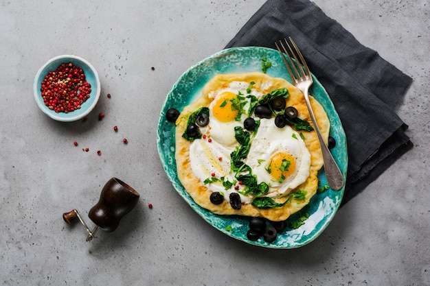 Türkisches Eierfladenbrot mit Joghurt, Käse, Oliven, Spinat und rotem Pfeffer auf keramischer Vintage-Platte auf grauer alter Oberfläche. Ansicht von oben.