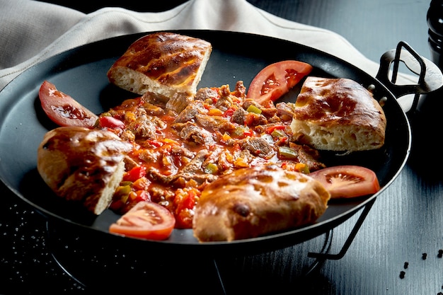 Türkisches Lammgulasch in roter Soße mit Paprika und traditionellen Brötchen in einer Metallschale auf einem schwarzen Tisch. Nahaufnahme, selektiver Fokus