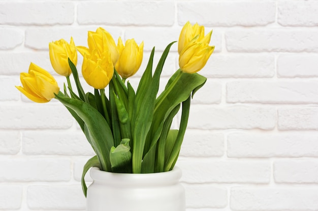 Tulipanes amarillos en florero. Flores florecientes de la primavera brillante en la pared de ladrillo decorativa blanca. Florido natural.