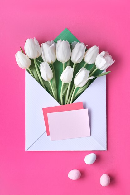 Tulipanes blancos en sobres de papel con tarjetas en blanco y huevos de Pascua sobre fondo rosa vibrante.