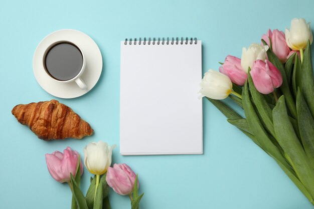 Tulipanes, cuaderno en blanco, croissant y taza de café sobre fondo azul.