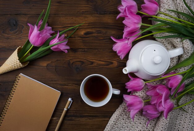 Los tulipanes, un cuaderno, una tetera blanca y una taza de té se colocan sobre una mesa de madera