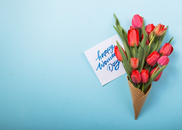 Tulipanes rojos hermosos en un cono de galleta de helado con tarjeta Día de la mujer sobre un fondo concreto. Idea conceptual de un regalo de flores. Humor de primavera