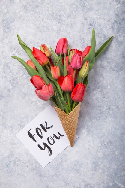 Tulipanes rojos hermosos en un cono de galleta de helado con tarjeta para usted sobre un fondo concreto. Idea conceptual de un regalo de flores. Humor de primavera