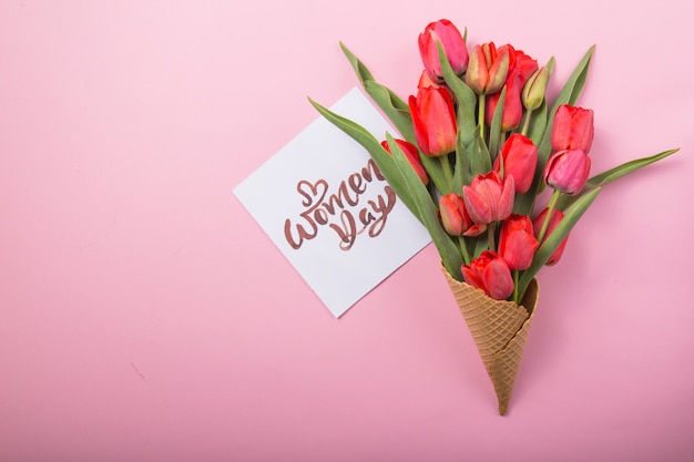 Tulipanes rojos hermosos en un cono de galleta de helado con tarjeta Womans Day sobre un fondo de color. Idea conceptual de un regalo de flores. Humor de primavera