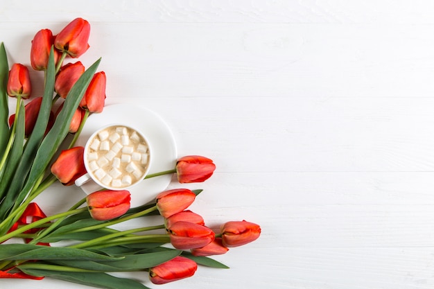 Tulipanes rojos y una taza de café con malvaviscos en blanco. Preparando una postal para las vacaciones de primavera. Copia espacio