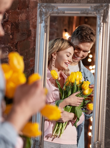 Tulipas amarelas. Mulher bonita radiante segurando tulipas amarelas ao lado do marido e olhando no espelho