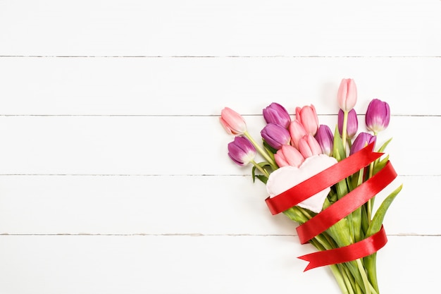 Tulipas com coração vermelho da fita e da tela no fundo de madeira branco.