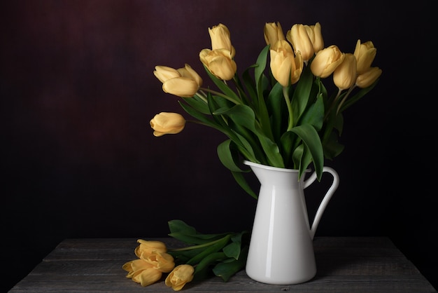 Tulpen in einem Krug. Klassisches Stillleben mit einem Strauß gelber Tulpenblumen in einem weißen Weinlesekrug auf dunklem Hintergrund und einem alten Holztisch.