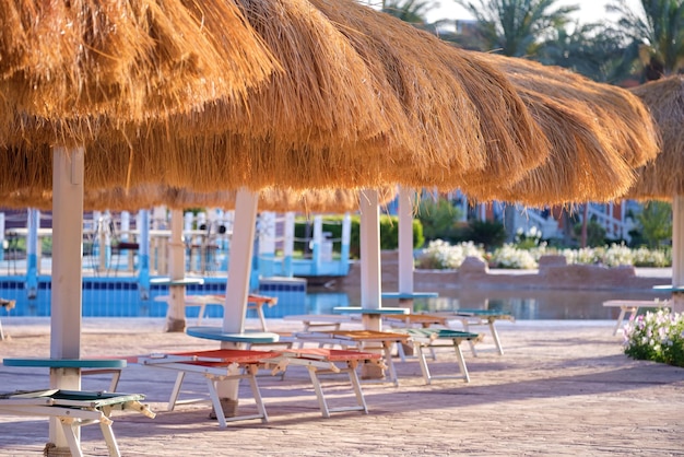 Tumbonas vacías bajo sombrillas de paja en el lado de la piscina en un complejo tropical Vacaciones de verano y concepto de escapada