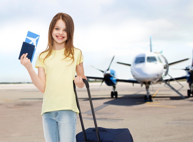 turismo, férias, infância e conceito de transporte - menina sorridente com mala de viagem, bilhete e passaporte sobre o fundo do aeroporto