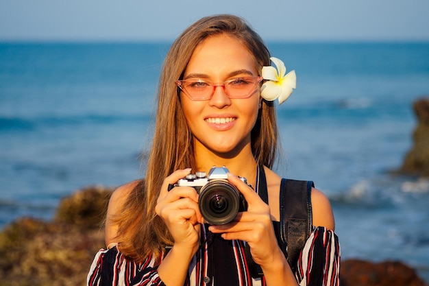 Turismo tropical de verão e fotografia. jovem mulher viajando em um elegante macacão listrado com câmera e mochila perto do mar, praia, oceano índico, goa, turquesa, água, céu, e, rochas.