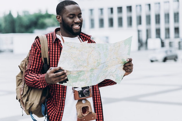 Turista afro-americano bonito e feliz com câmera antiga e mapa na cidade moderna