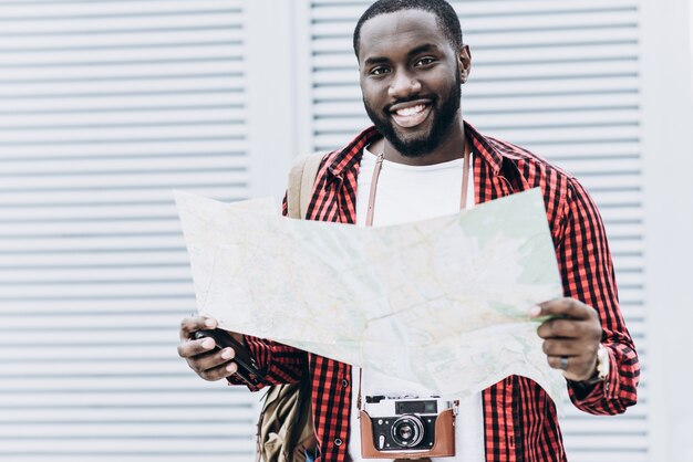 Turista afro-americano bonito e feliz com câmera antiga e mapa na cidade moderna