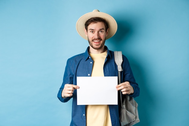 Turista alegre com chapéu de verão, mostrando o pedaço de papel vazio e sorrindo, pegando carona com a mochila, de pé sobre fundo azul.