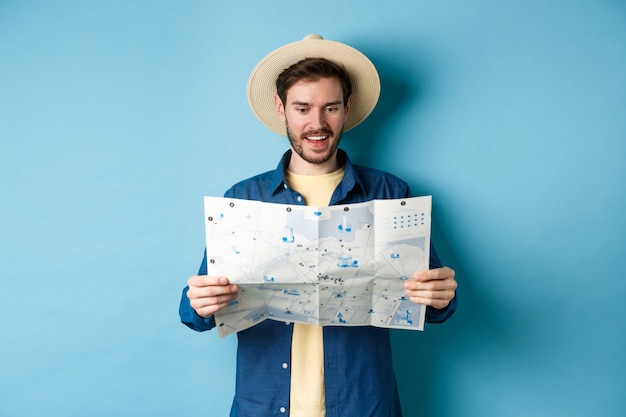 Turista alegre en ruta de viaje de planificación de sombrero de verano en vacaciones, mirando el mapa de turismo y sonriendo emocionado, de pie sobre fondo azul.