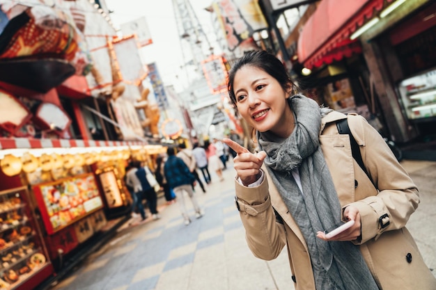 turista alegre sosteniendo un mapa en línea y buscando el restaurante para almorzar en el izakaya local japonés. joven viajera señalando alegremente con el dedo al destino en la bulliciosa calle osaka, japón.