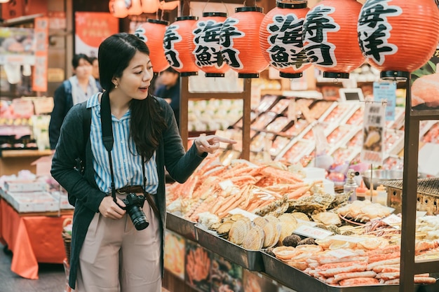 turista asiática apontando para frutos do mar frescos exibidos em uma loja que vende produtos aquáticos no mercado kuromon ichiba em Osaka, Japão. tradução na lanterna: â€œfrutosâ€ e âsushiâ