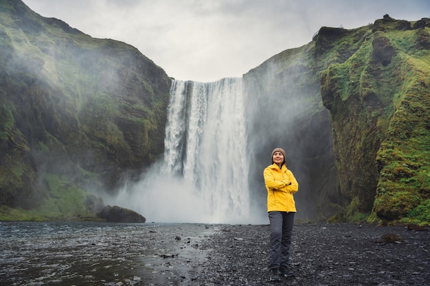 Turista asiática de jaqueta amarela apreciando a cachoeira Skogafoss fluindo do penhasco no verão na Islândia