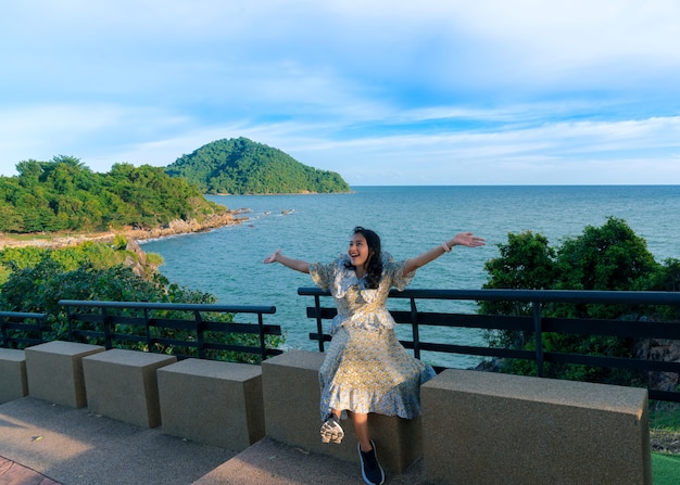 Turista asiática se senta e se sente revigorada com o braço feliz erguido no assento ao lado da passarela no mirante Noen Nang Phaya com vista para o mar azul e a montanha, atração turística em Chanthaburi, Tailândia