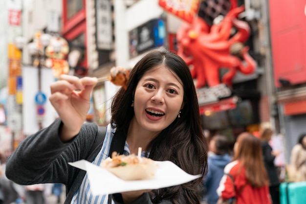 turista asiática sorridente segurando mostrando uma bola de takoyaki na picareta e olhando para a câmera enquanto visitava shinsaibashi suji e doutonbori no centro de Osaka, japão