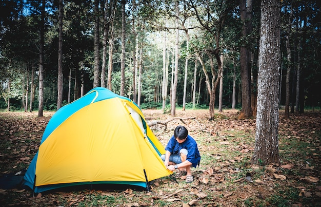 Un turista asiático, lanza una carpa amarilla para acampar en un bosque con árboles altos, a las personas y al concepto de viaje.