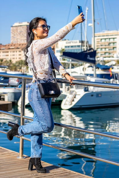 Turista asiático sonriente se toma un selfie con su teléfono móvil en el puerto con barcos en el fondo