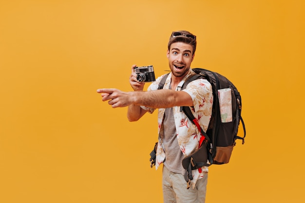 Turista barbudo com olhos bonitos em uma roupa descolada de verão, olhando para a câmera e posando com uma mochila grande na parede isolada