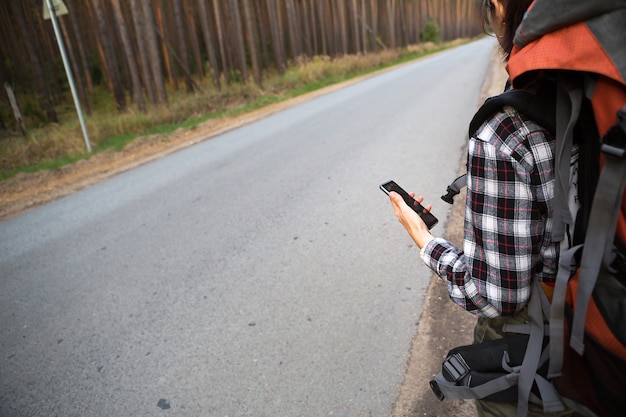 Turista en una camisa a cuadros con una mochila grande naranja cerca de una carretera en el bosque con un teléfono inteligente en la mano. Navegación, mapas satelitales, comunicación, turismo interno. Mochilero, aventura