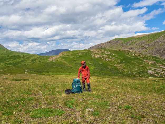 Turista cansada com uma grande mochila e bolsas, felizmente, olha para longe no contexto das nuvens dramáticas azuis no contexto das montanhas de tundra.