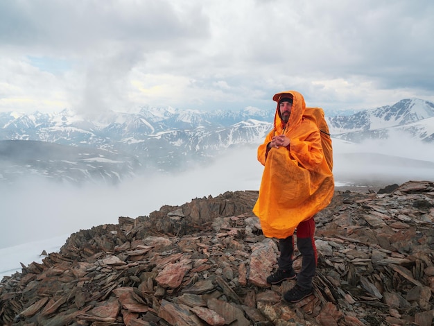 Turista cansado con un impermeable amarillo en la cima de una montaña se mira a sí mismo y se refleja en el telón de fondo de nubes dramáticas. Senderismo en solitario con una mochila grande en la montaña.