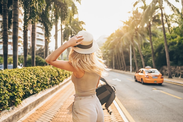 Turista chinês andando nas ruas da cidade no turismo de férias na Ásia. Mulher bonita caucasiana no chapéu com mochila viajar conceito de estilo de vida. Rua andando, palmeiras verdes e rodovia em Sanya, Hainan.