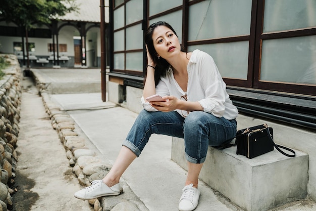 turista chinesa asiática usando telefone celular aproveitando as férias de viagem de verão. jovem visitando a cidade velha em kyoto, japão. linda fêmea sentada na cadeira de banco de pedra com celular e bolsa.