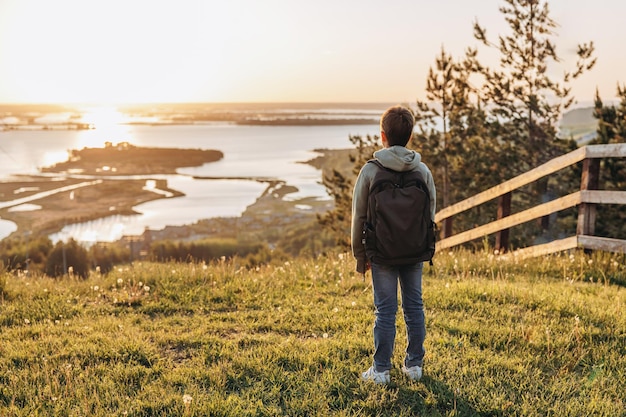 Turista com mochila de pé no topo da colina no campo de grama e apreciando a bela vista da paisagem Vista traseira do alpinista adolescente descansando na natureza Estilo de vida ativo Conceito de viagens locais