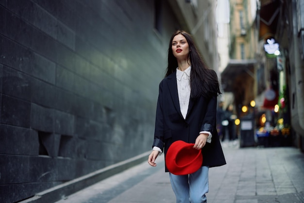 Turista da moda em roupas elegantes em uma jaqueta e boina vermelha andando por uma rua estreita da cidade cabelo voador e viagem feliz Estilo francês cor cinematográfica estilo retrô